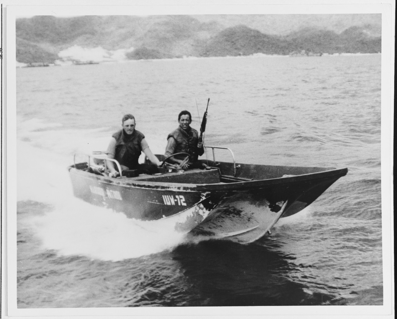 Black-and-white historical photograph of two men traveling in a U.S. Navy Boston Whaler skimmer in Da Nang Harbor in 1966.